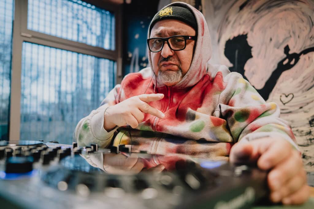 DJ Oldschool Peppino a keverőpult mögött. Idősebb, szemüveges, őszülő szakállú férfi fekete kötött sapkában, amire a drapp-zöld-piros foltmintás felsőjének kapucniját is ráhúzta. Jobb kezével V betűt, a győzelem jelét mutatja.