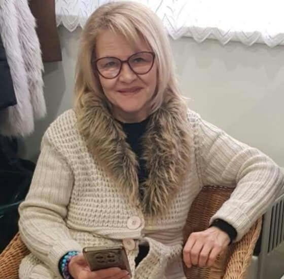 Szőke, félhosszú hajú, szemüveges, mosolygós nő fekete pulcsi és drapp, a nyaka körül műszőrmés kötöttkabát van rajta,