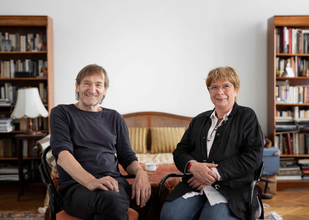 Cserhalmi György és Csáki Judit félig egymás felé fordított karosszékekben ülnek, mosolyognak. A hattérben középen üres, fehér falfelület, a két szélen két nagy, barna könyvesszekrény.