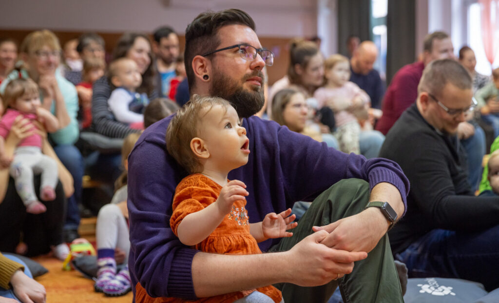 a kép középpontjában egy fiatal szakállas, szemüveges férfi van, ölében egy babával. Körülöttük, a háttérben is szülők láthatók, ölükben gyerekekkel. Mind egy irányba, jobbra figyelnek, vélhetőleg valamilyen előadásra, vagy előadóra.