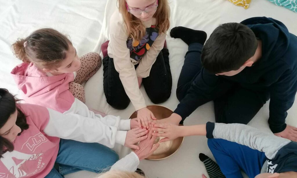 10 év körüli gyerekek ülnek körben a földön, egy fehér takarón. Egyik kezüket mind befelé, középre nyújtják, egy réz hangtál fölé.