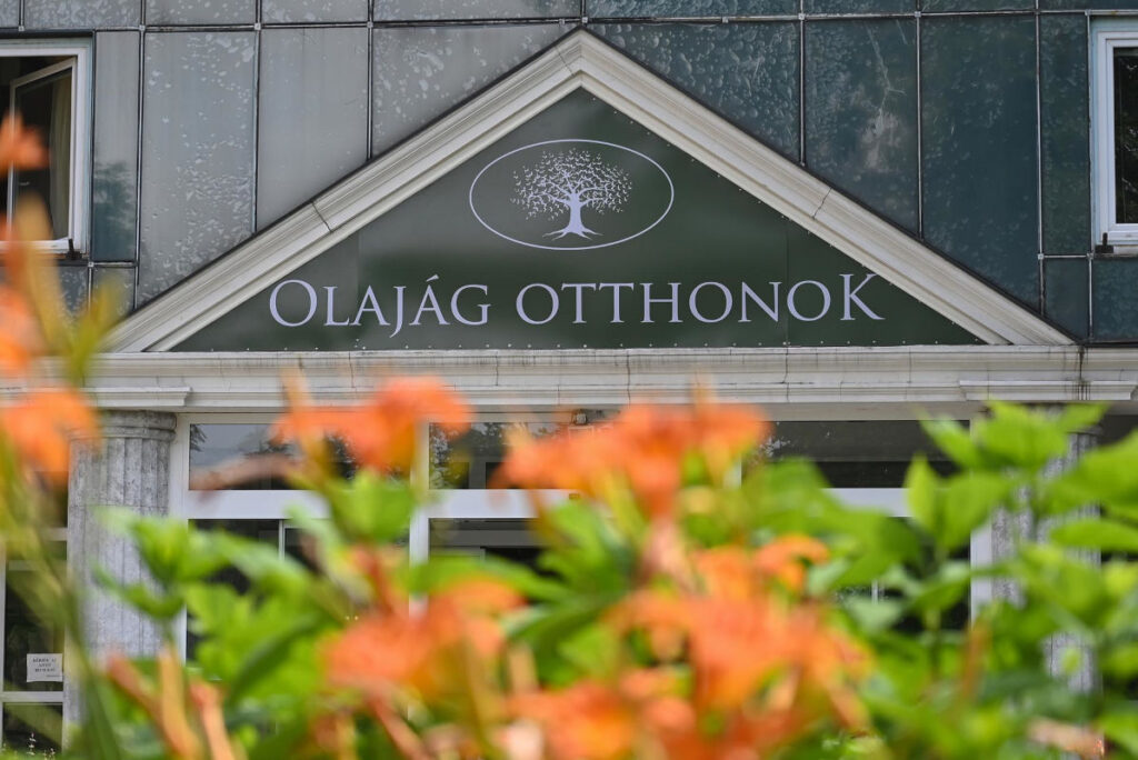 Az Olajág Otthonok épületének bejárata látható. A bejárat fölött az „OLAJÁG OTTHONOK” felirat és a logó szerepel. Az előtérben elmosódott narancssárga virágok és zöld növények láthatók, a háttérben az épület homlokzata és üvegfelületei jelennek meg.