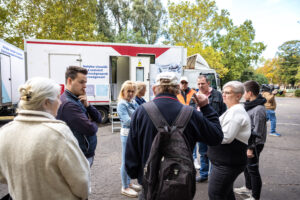 A közösségi szűrőprogram résztvevői és a rendezvényre érkező önkormányzati, valamint egészségügyi vezetők beszélgetnek a Nemzedékek parkjában felállított szűrőkamion előtt.