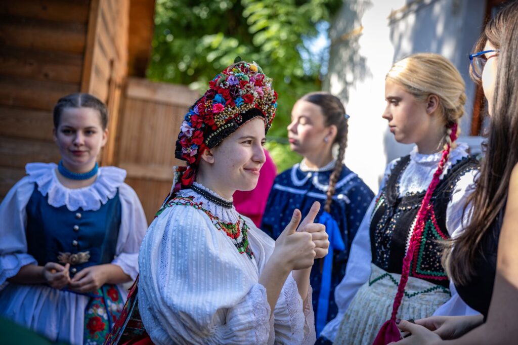 A Fóti Népművészeti Szakgimnázium és Gimnázium növendékei népviseletben készülnek a bemutatóra a XV. Kerületi Kulturális Örökség Háza és Helytörténeti Gyűjtemény udvarán. Előtérben egy gazdagon díszített, színes virágokkal ékesített fejdíszt viselő résztvevő látható, körülötte hagyományos viseletet öltött fiatalok beszélgetnek a program kezdete előtt.