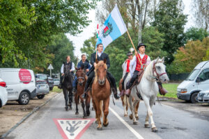 A rákospalotai szüreti felvonulás lovas résztvevői haladnak végig az utcán. Az élen haladó lovas a XV. kerület kék-fehér zászlaját viszi, mögötte további lovasok követik a menetet hagyományos ünnepi öltözetben.