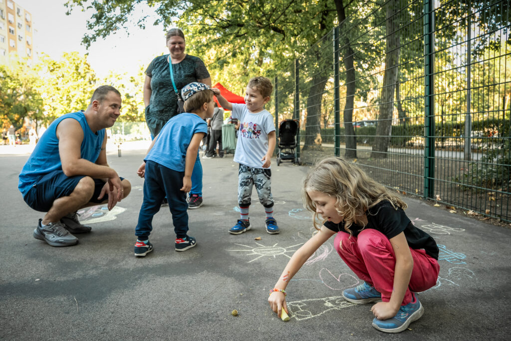 A szomszédünnepen a gyerekek krétarajzokat készítenek az aszfalton, miközben a családok a sportpályán szervezett programokon vesznek részt.