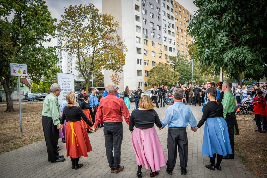 A SpeciArt Speciális Művészeti Fesztivál résztvevői egy körben állva, egymás kezét fogva adnak elő nyitótáncot a szabadban. A háttérben nézők, fotósok és lakóházak láthatók az Újpalotai Közösségi Ház környezetében.