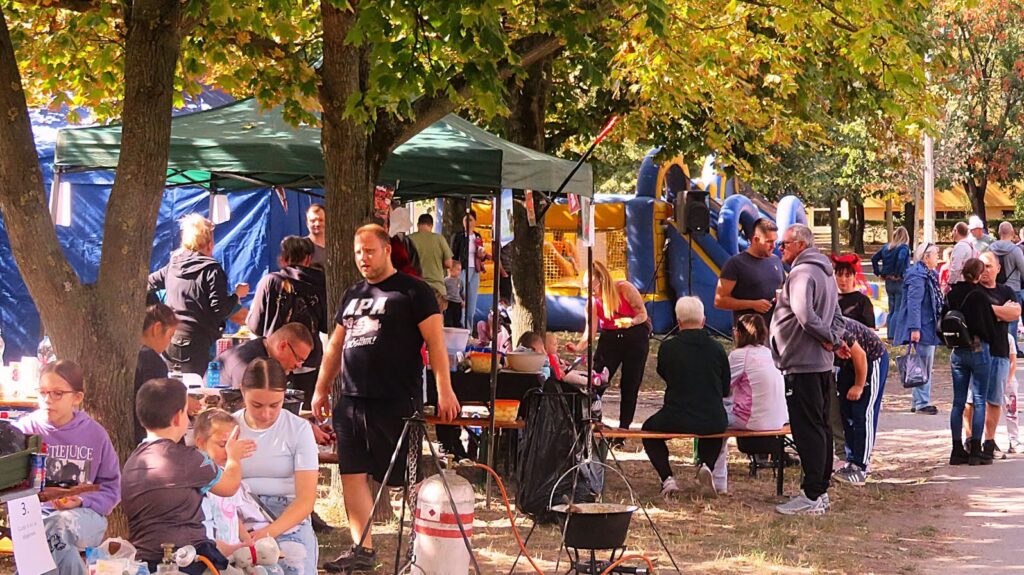 A Csobogós lakótelepen megrendezett szomszédünnepen a fák árnyékában felállított sátrak, asztalok és padok körül beszélgetnek a résztvevők. A háttérben ugrálóvár látható, az előtérben pedig közös főzés és közösségi programok zajlanak. A rendezvény családokat és környékbeli lakókat hozott össze egy kötetlen, szabadtéri közösségi napon.