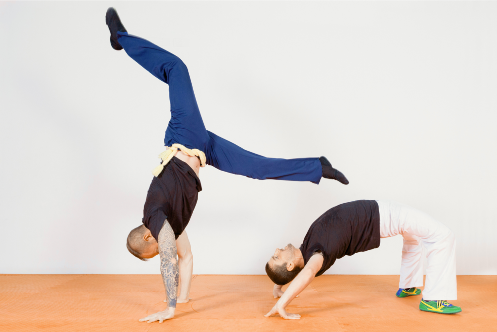 Egy fehér falú teremben két férfi capoeirázik. A jobb oldali férfi hídba van, a bal oldali pedig kézen áll, lábait haránt terpeszben tartva.