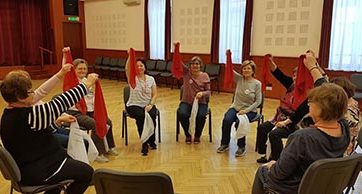Egy világos teremben idősebb hölgyek ülnek körben, székeken. Vidáman mosolyognak, és élénkpiros kendőket tartanak a magasba.
