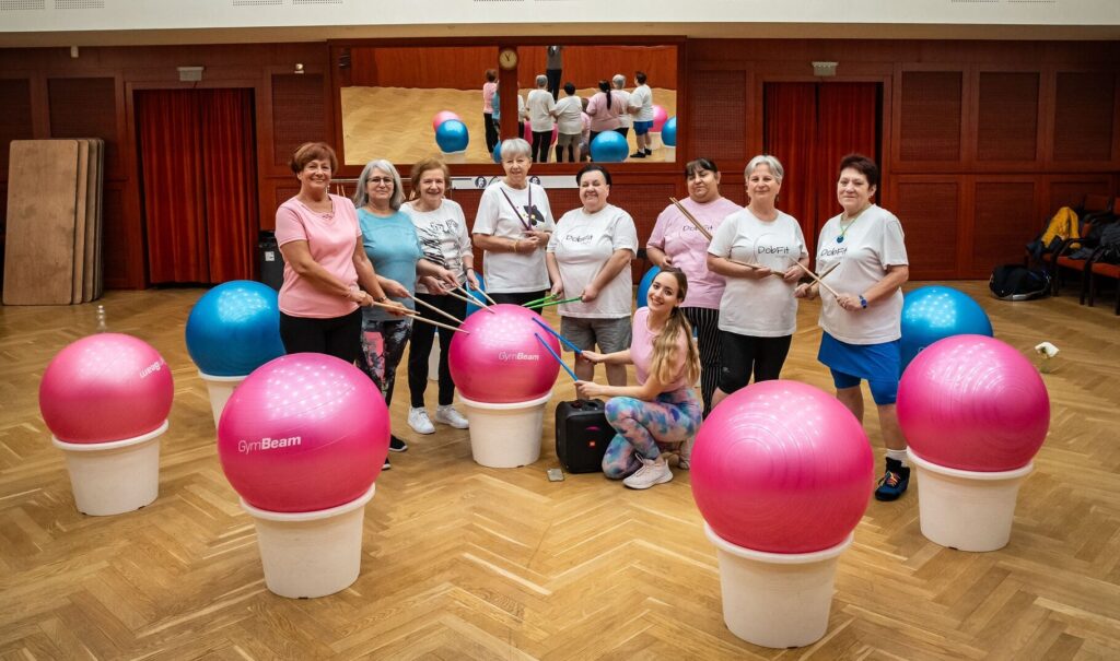 Csoportkép néhány dobfitesről és az edzőjükről. A kezükben dobverő, előttük, körülöttük nagy világoskék és pink sportlabdák, amik az edzés során dobként szolgálnak.