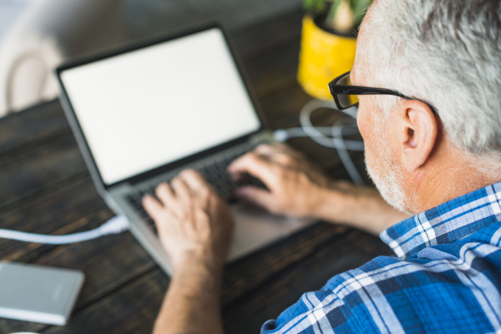 Egy ősz hajú, szemüveges, kék skótkockás inget viselő férfi bal válla fölött látunk a képre. Mindkét kezével ír egy kisméretű laptopon, ami egy rusztikus, sötétbarna lécekből álló asztalon van.