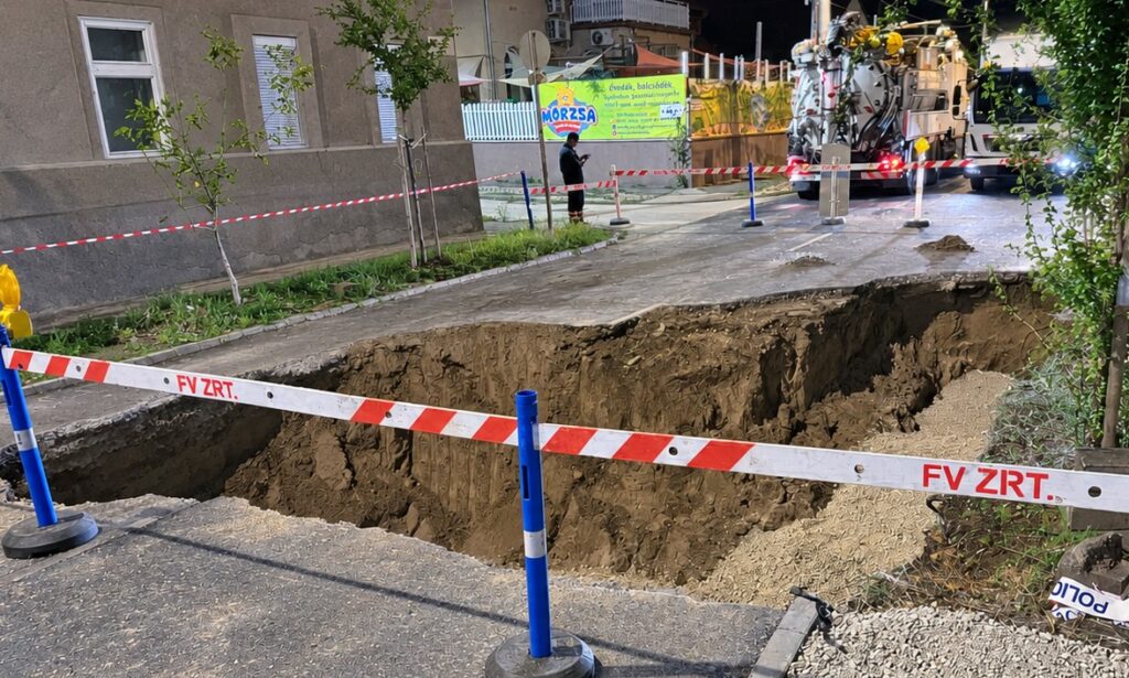 Egy nagy kiterjedésű útszakadás látható a Sződliget utcában éjszaka. Az úttest közepén mély gödör keletkezett, amelyet piros-fehér szalaggal és kék terelőoszlopokkal kerítettek körbe. A háttérben egy munkagép és egy utcai házsor látható.