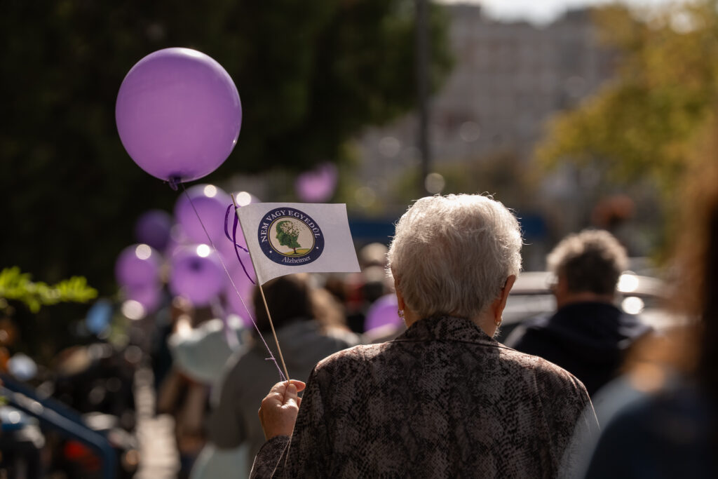 Idős résztvevő egy szabadtéri rendezvényen lila léggömböt és az Alzheimer-betegségre figyelmet felhívó zászlócskát tart a kezében. A háttérben több ember és további lila léggömbök láthatók.