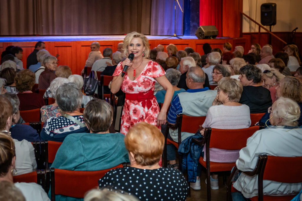 Szendy Szilvi mikrofont tartva énekel a Csokonai Rendezvényház nézőtere előtt az Idősek Világnapja alkalmából rendezett ünnepségen. A művész a széksorok között halad, miközben a közönség figyelemmel kíséri az előadást.