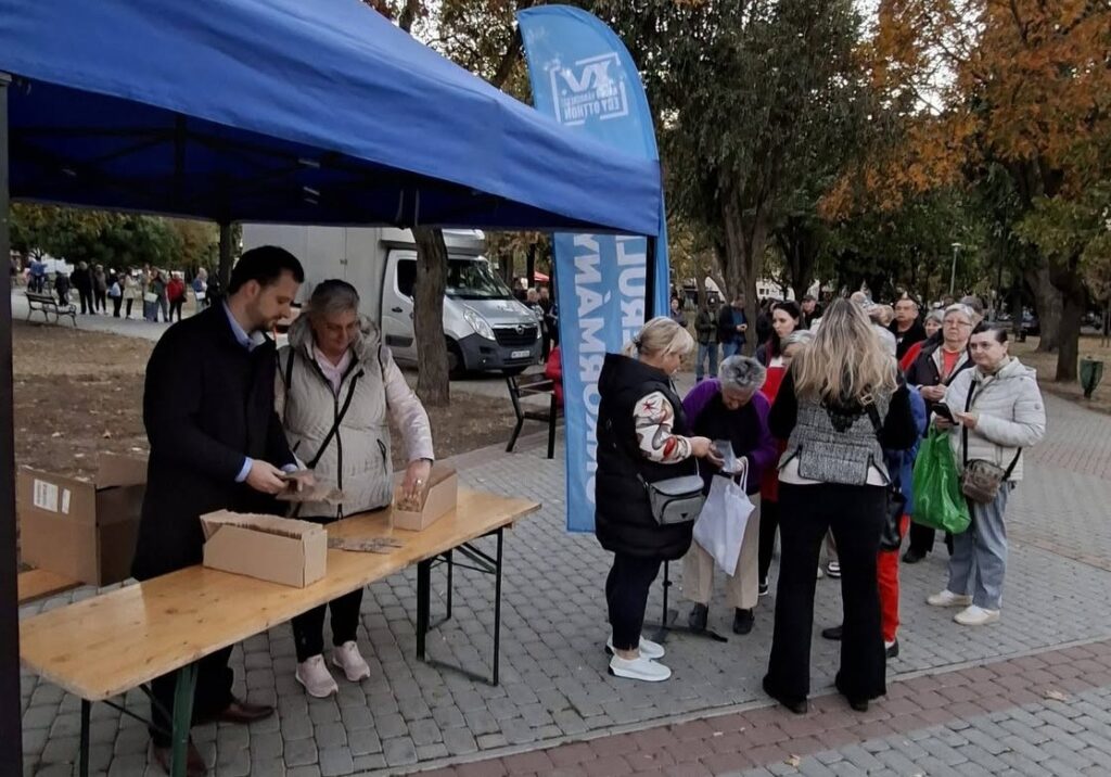 A virágmagosztáson az asztal mögött álló önkormányzati vezetők adják át a vadvirágmag-keveréket az érdeklődőknek, miközben a résztvevők sorban várják a csomagok átvételét. Középen Cserdiné Németh Angéla polgármester és Gyurkó Dániel alpolgármester látható.