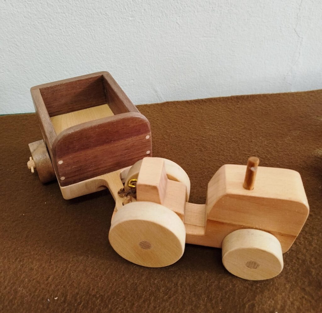 Fából készült, a kép jobb oldala felé "haladó" natúr színű kis traktor, mögötte bekanyarodó utánfutóval. Alatta barna filc, a háttérben fehér fal.