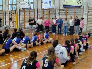 A XV. Szalayné Sebők Éva-emléktorna záróünnepségén a részt vevő csapatok játékosai a sportcsarnok pályáján ülve hallgatják a megemlékezés felszólalóit. A háttérben a torna és a Palota Röplabda SC molinói láthatók.