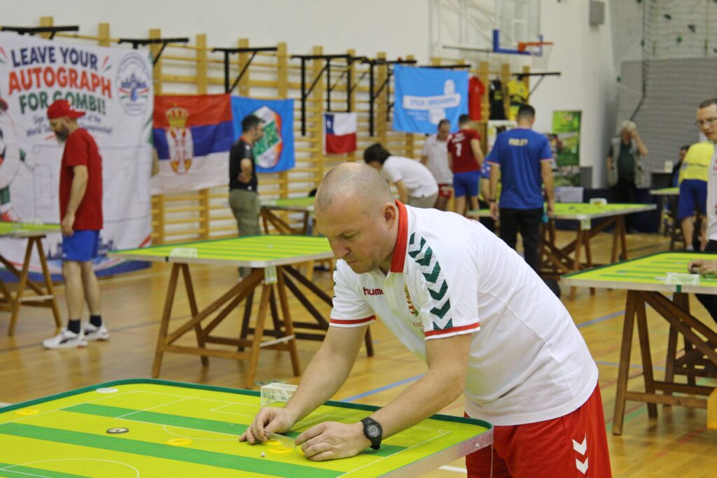 A budapesti szektorlabda-világbajnokság egyik résztvevője játék közben a Ludovika Sportcsarnokban. A háttérben több játékasztalnál zajlanak a mérkőzések, a csarnok falán a részt vevő országok zászlói láthatók.