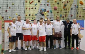 A magyar válogatott és a szakmai stáb tagjai a budapesti szektorlabda-világbajnokság eredményhirdetésén. A csapat tagjai érmeket viselnek, egyikük a világbajnoki trófeát emeli a magasba. A háttérben a verseny hivatalos molinói és a sportcsarnok mászófala látható.