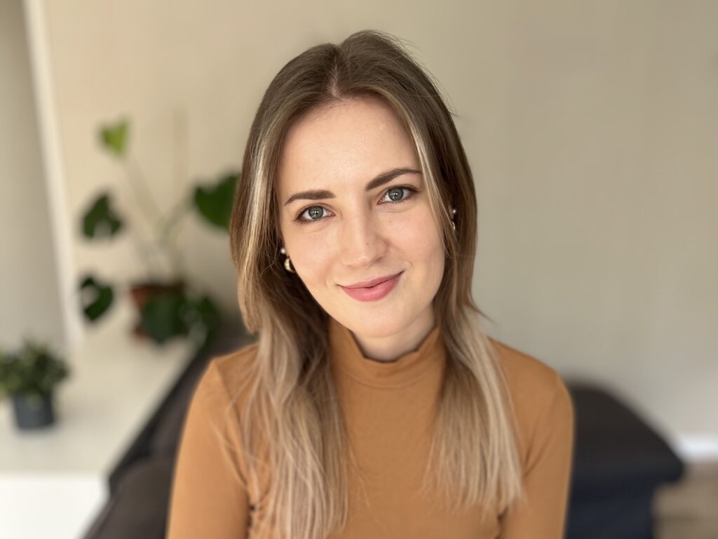 Mosolygós, sima félhosszú szőkésbarna hajú nő mellképe, kissé felfelé néz, frlülről lett fotózva. A szeme szürkéskék. Őzbarna vékony garbót visel. A kép baloldalán a háttérben elmosódva két cserepes növény látható.