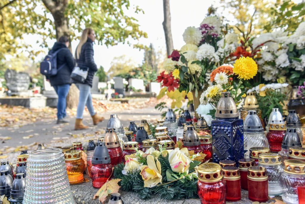 Virágokkal és mécsesekkel díszített síremlék egy temetőben. Az előtérben különböző színű kegyeleti gyertyák és virágcsokrok láthatók, a háttérben pedig látogatók sétálnak a sírok között az őszi lombhullás idején.